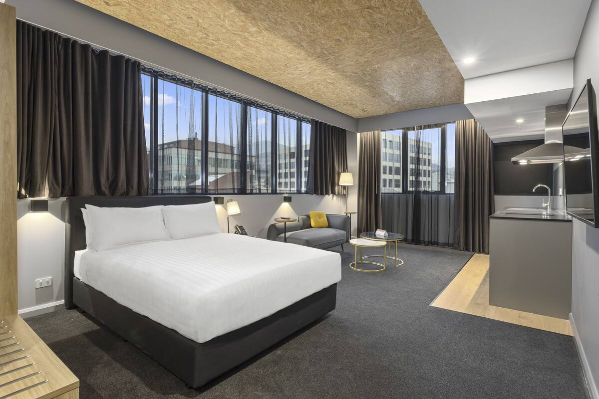 A modern hotel room with a large bed, a sofa, a small table, and a kitchenette.