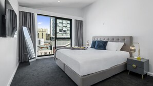 Una habitación de hotel moderna con una cama grande, una mesita de noche con lámpara y vistas a la ciudad a través de amplios ventanales.