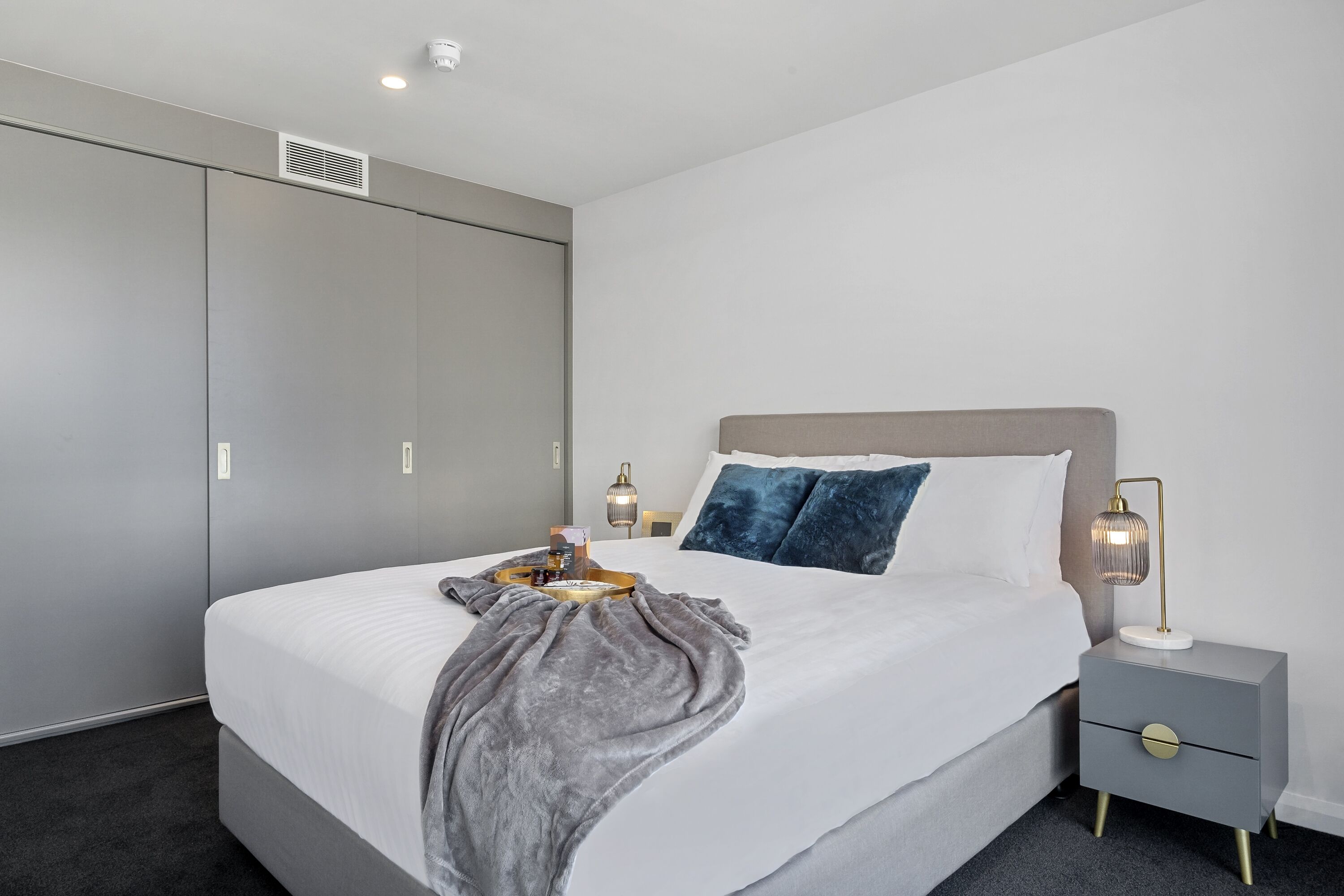 Un dormitorio moderno con una cama grande, un armario gris, lámparas de noche y una bandeja con objetos sobre la cama.