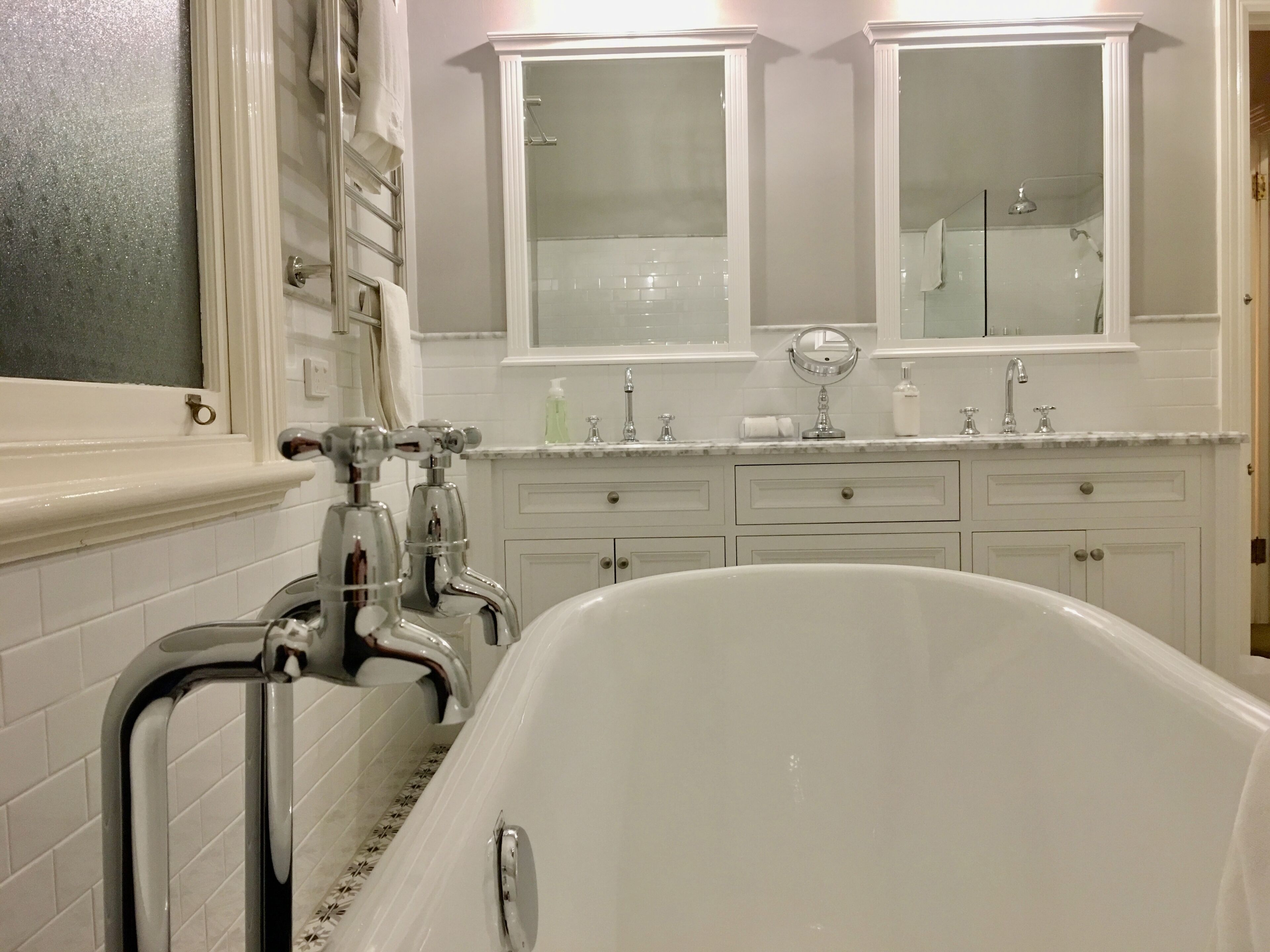 A bathroom with a freestanding bathtub, white tiled walls, a double vanity with two mirrors, and a glass shower enclosure.