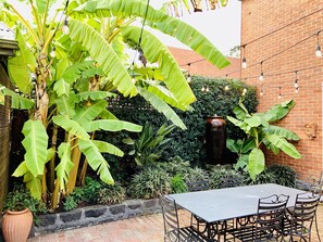 Une terrasse en briques avec une table et des chaises, entourée de bananiers luxuriants et d’un mur en briques.