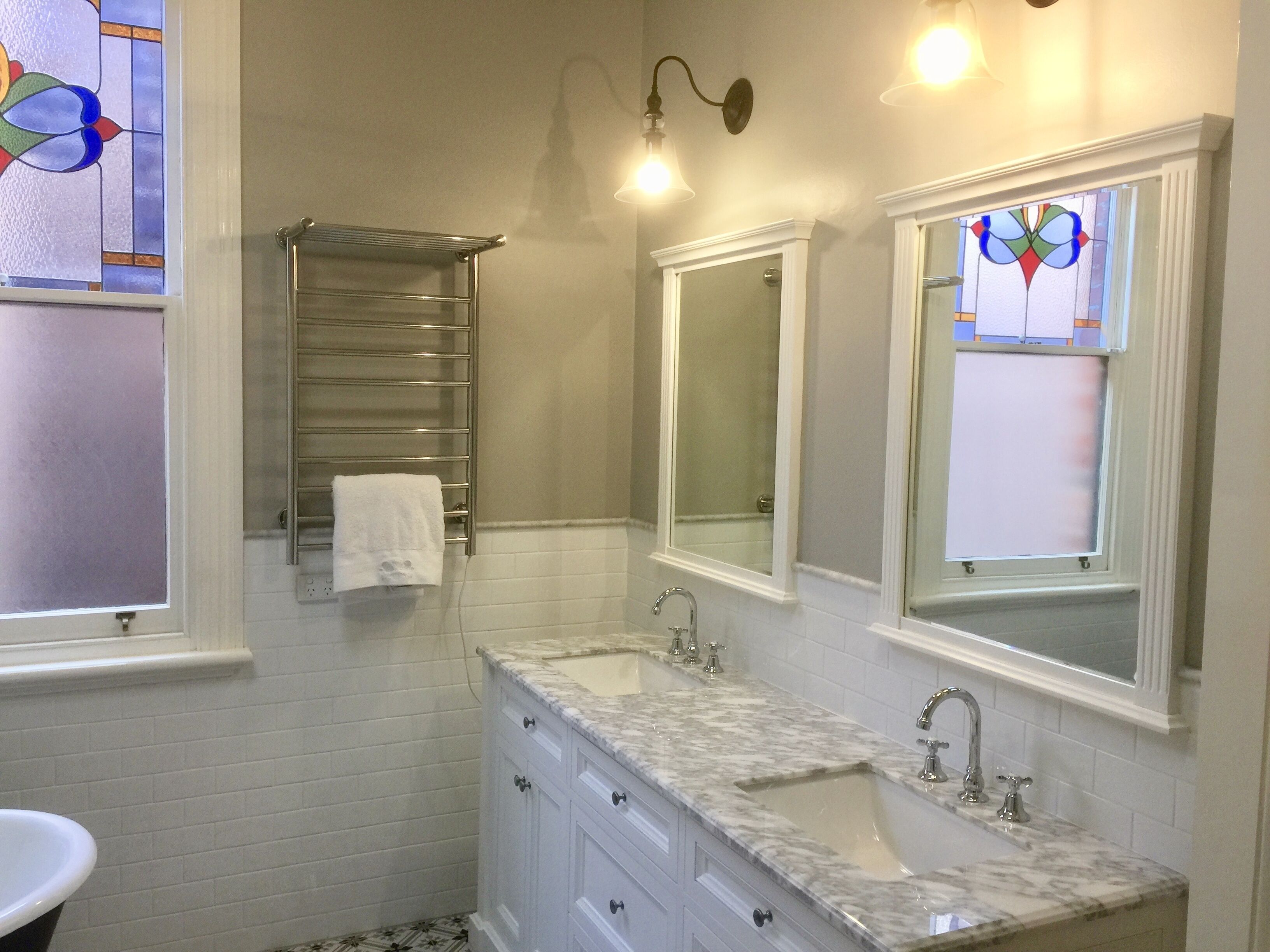 A bathroom with two sinks, a mirror, a towel rack, and a window with stained glass.