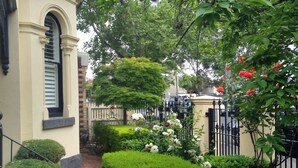 Un jardín cuidado con diversas plantas y flores, un sendero de ladrillo y un edificio tradicional con una ventana y una puerta.