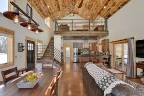 A spacious loft-style living area with a high ceiling, exposed wooden beams, a dining table with chairs, a sofa, and a kitchen area with a large island. - Riverfront Home w/ BBQ: 2 Mi to Downtown Gunnison! (Gunnison)