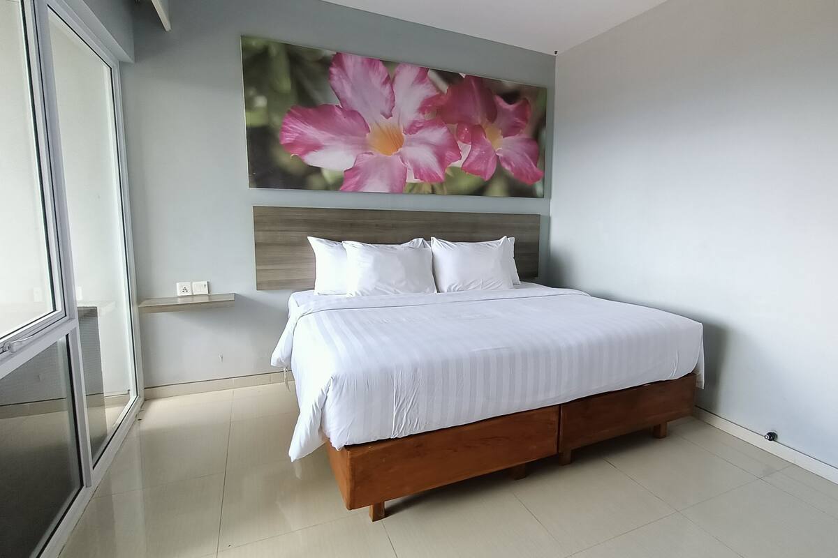 Une chambre à coucher avec un lit, du linge de lit blanc, une tête de lit en bois, un tableau représentant une fleur accroché au mur et une porte-fenêtre coulissante.