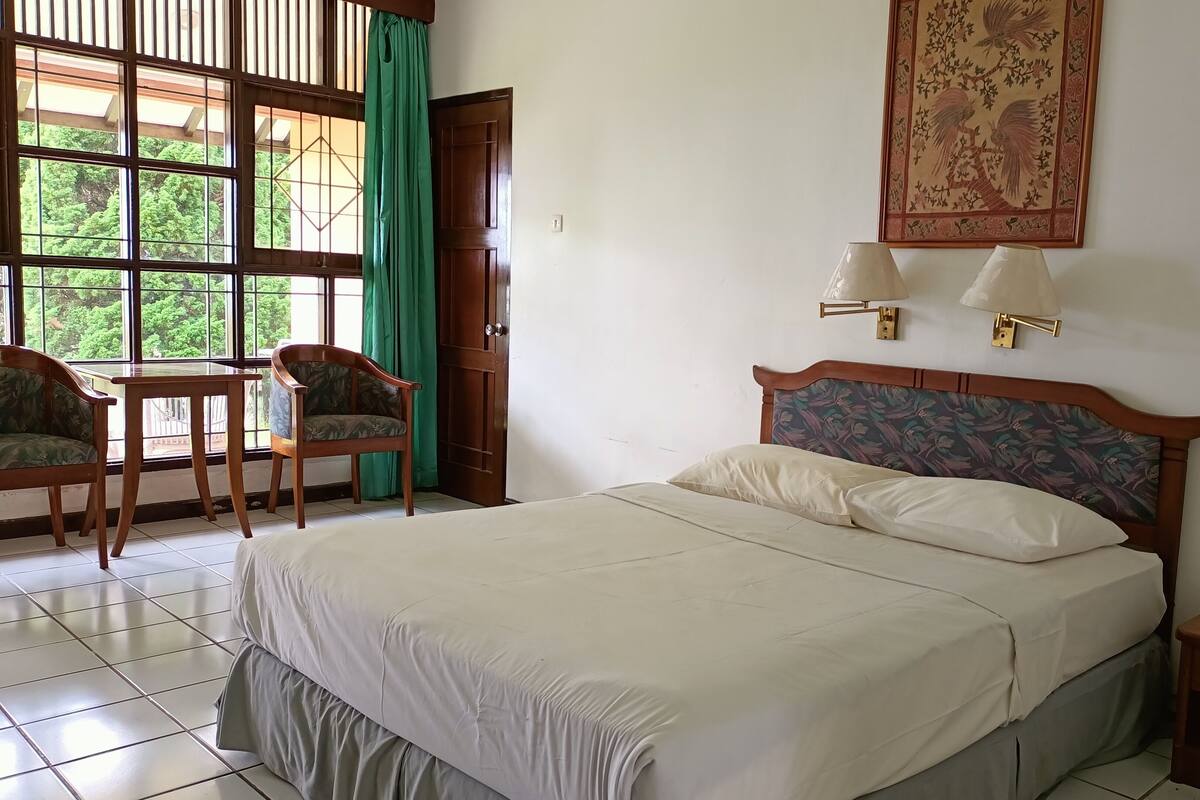 Une chambre d’hôtel comprenant un lit, deux chaises, une table, une fenêtre avec des rideaux et une lampe fixée au mur.