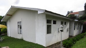 A small, single-story house with a white exterior, a brick pathway leading to the front door, and a small garden area with grass and shrubs. - Amanda Hills Bandungan (Bandungan)