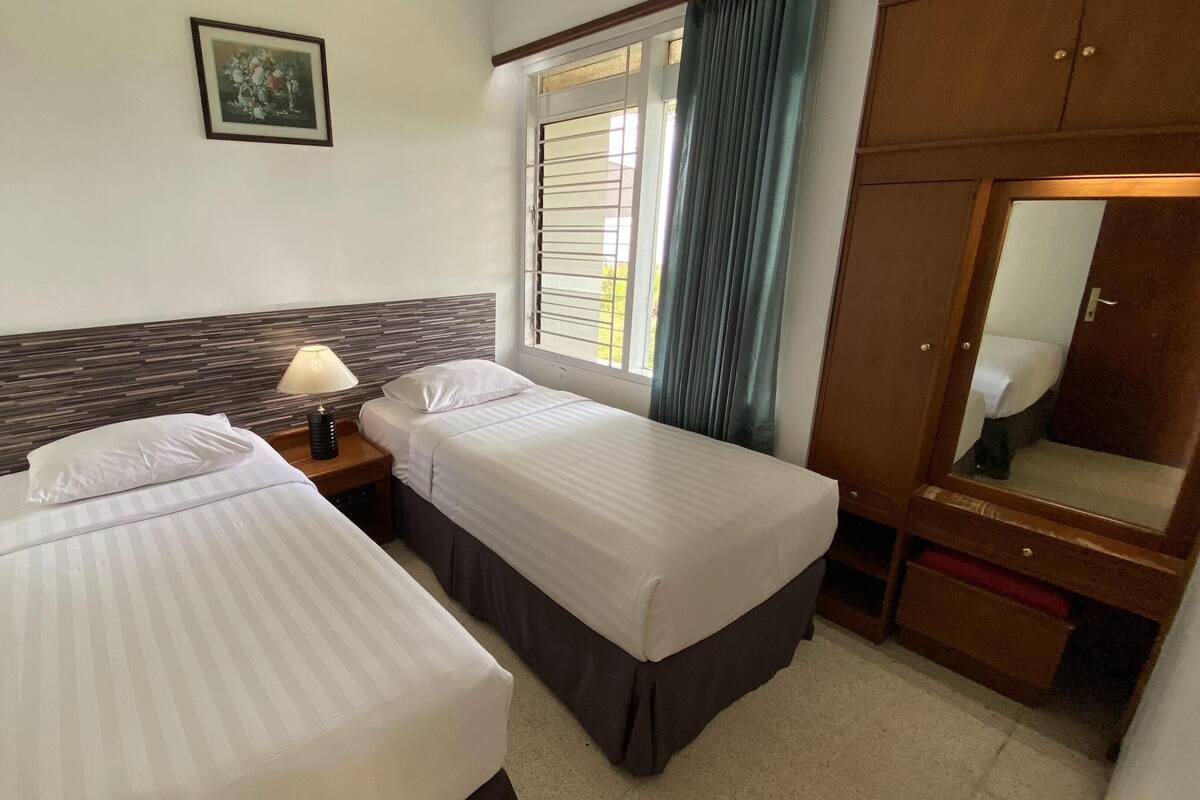 Une chambre d’hôtel avec deux lits simples, une table de chevet avec une lampe, une armoire en bois et une fenêtre avec des rideaux.