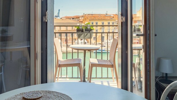 Een balkon met rieten stoelen en een tafel, met uitzicht op een stadsgezicht met gebouwen en een wateroppervlak.