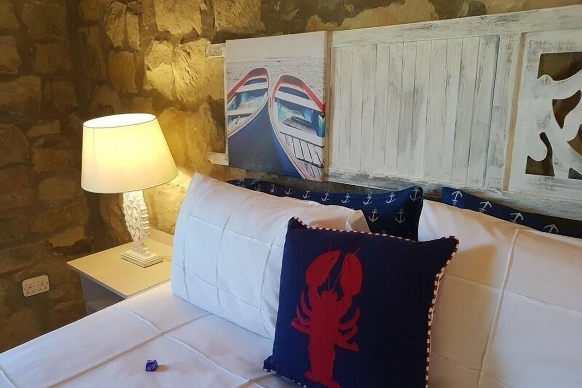 Une chambre avec un mur en pierre, un lit blanc avec un oreiller bleu marine orné d’un motif représentant un homard rouge, une table de chevet blanche avec une lampe, et un tableau encadré représentant des bateaux accroché au mur.