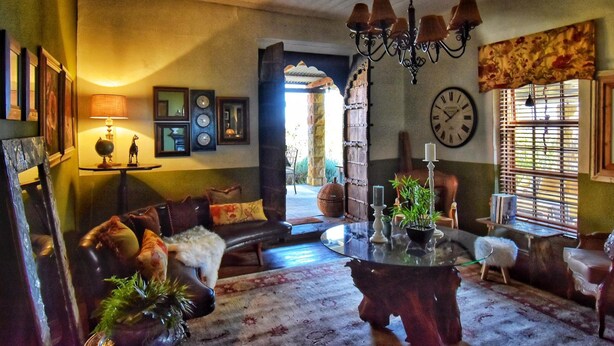 A cozy living room with a leather sofa, a wooden coffee table, and a chandelier. There is a clock on the wall and a window with blinds. - Affi Lande Boetiek Guest Farm (Fouriesburg)
