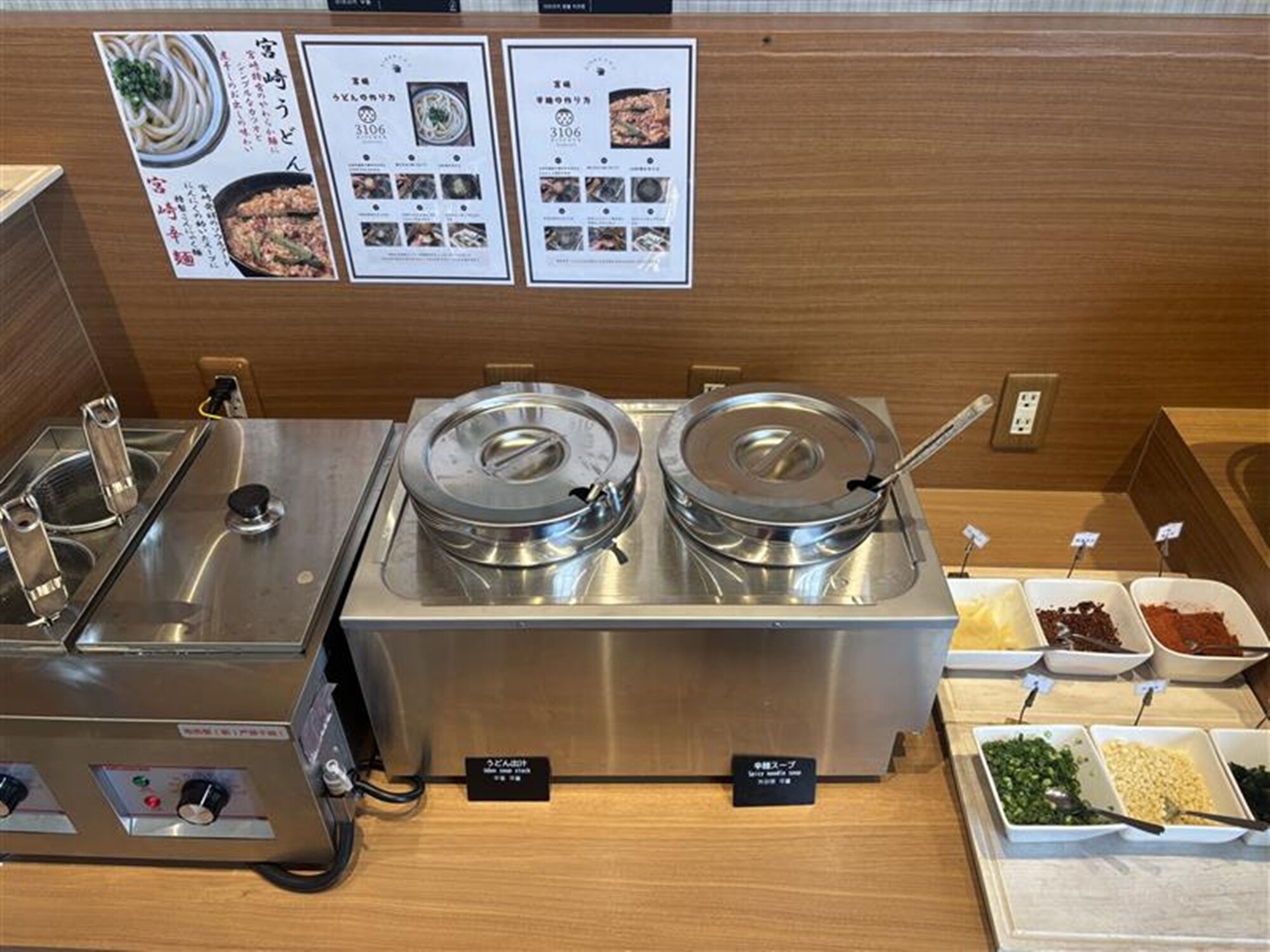 A buffet setup with two stainless steel chafing dishes, a variety of condiments, and a menu board above.