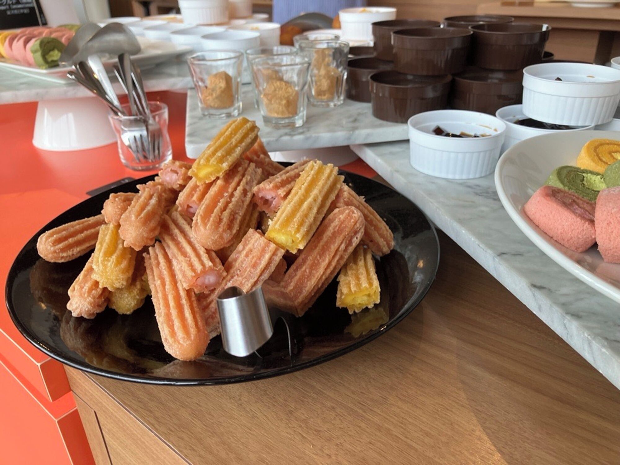 Un plato de churros con una cuchara de metal para servir, sobre un plato negro, rodeado de pequeñas tazas y recipientes de salsa de chocolate.