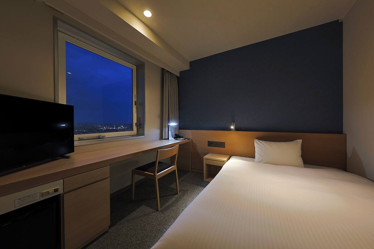 Une chambre d’hôtel comprenant un lit, un bureau avec une chaise, une télévision, une fenêtre donnant sur la ville de nuit et une table de chevet avec une lampe.
