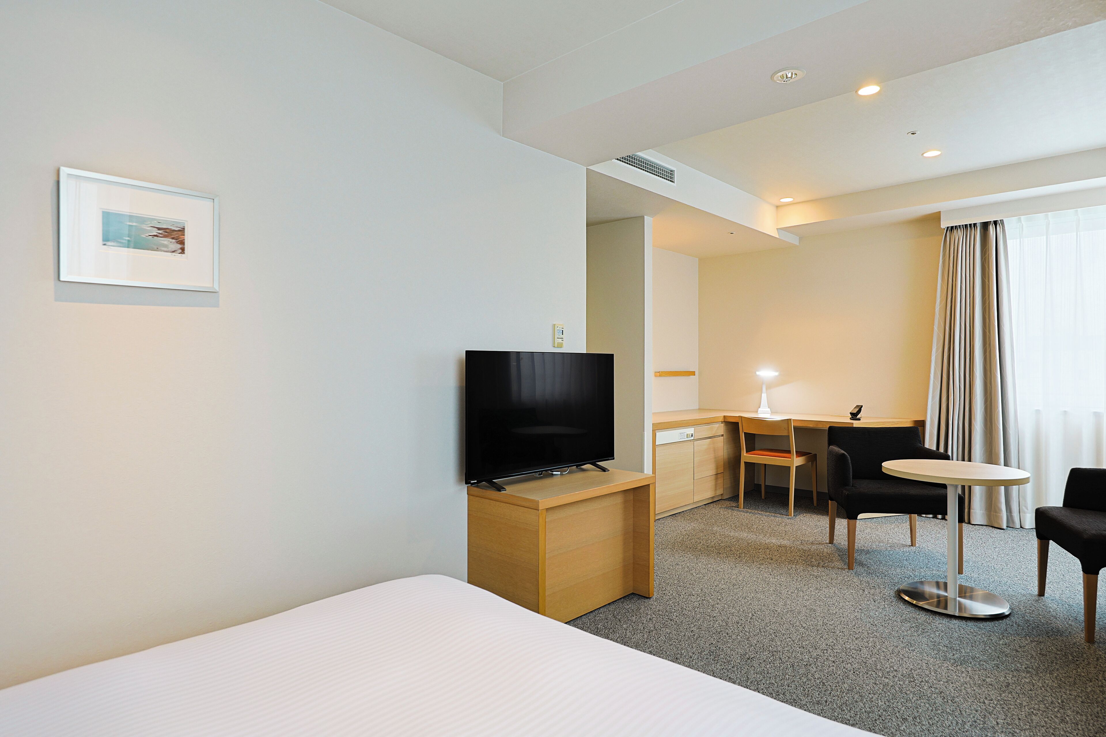 A hotel room with a bed, a TV on a wooden stand, a desk with a chair, and a small round table.