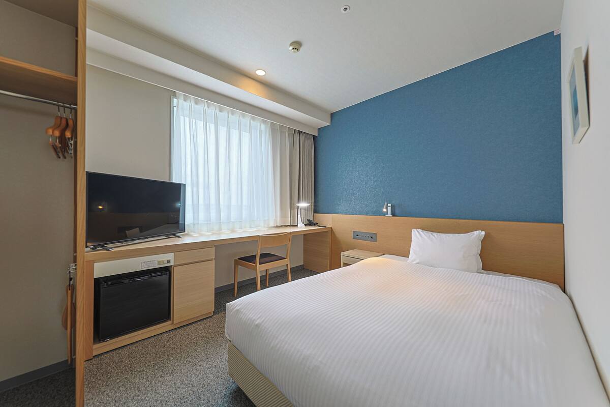 Une chambre d’hôtel comprenant un lit, un bureau avec une télévision, une chaise, une petite table de chevet et une fenêtre avec des rideaux.