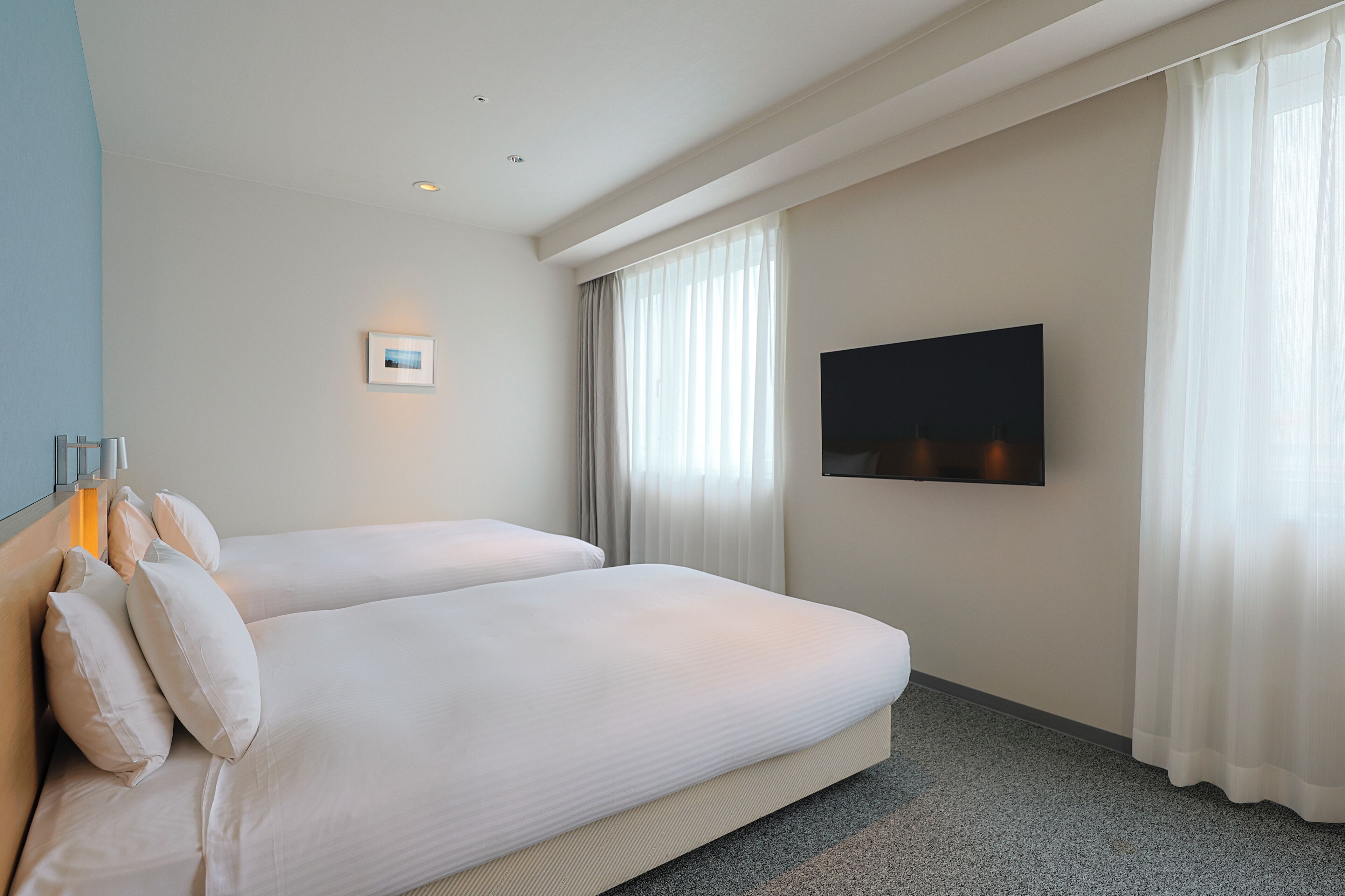 A hotel room with two beds, a flat-screen TV mounted on the wall, and a window with sheer curtains.