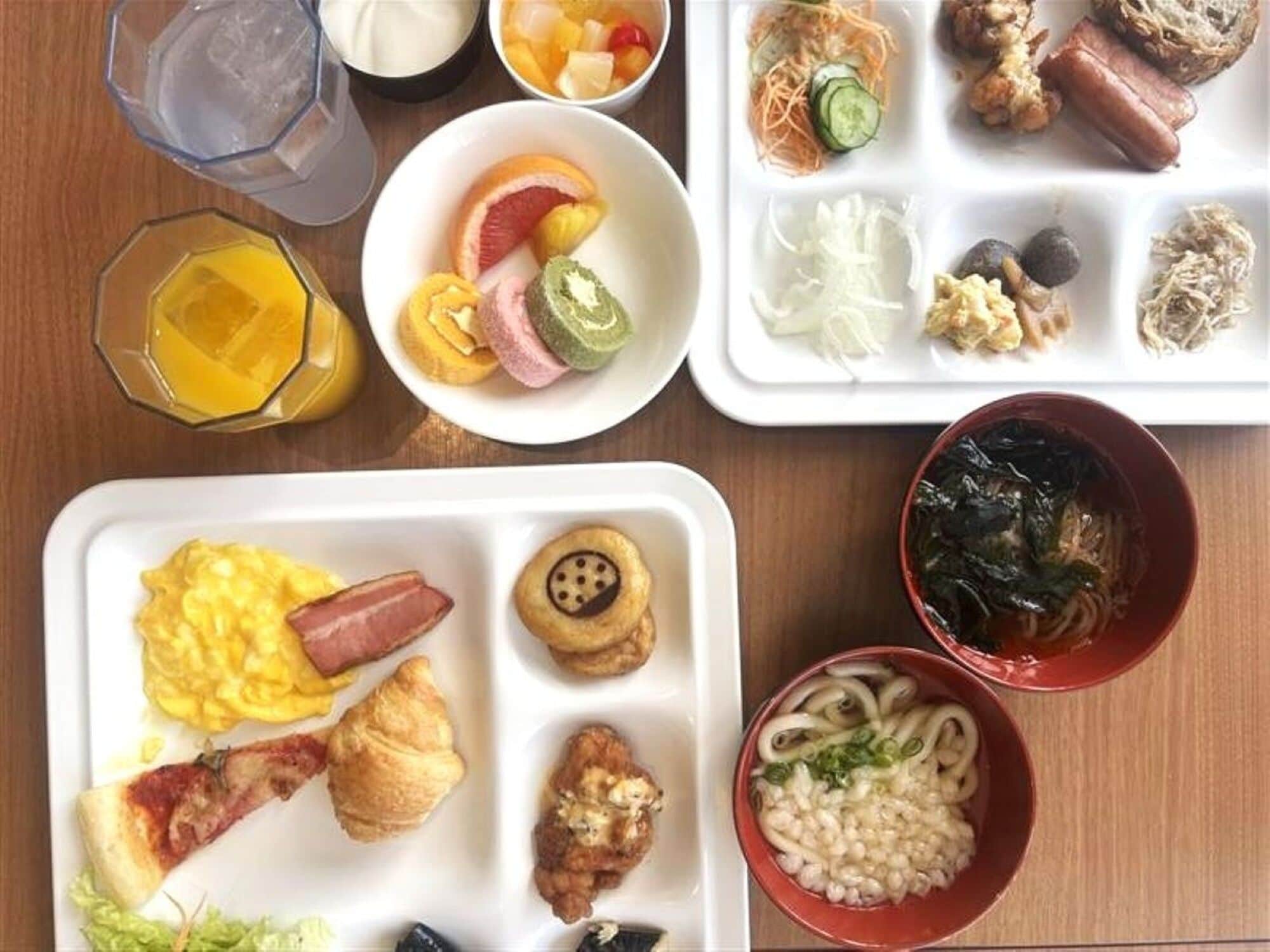 A breakfast spread with various dishes including eggs, bacon, croissants, and a bowl of noodles.
