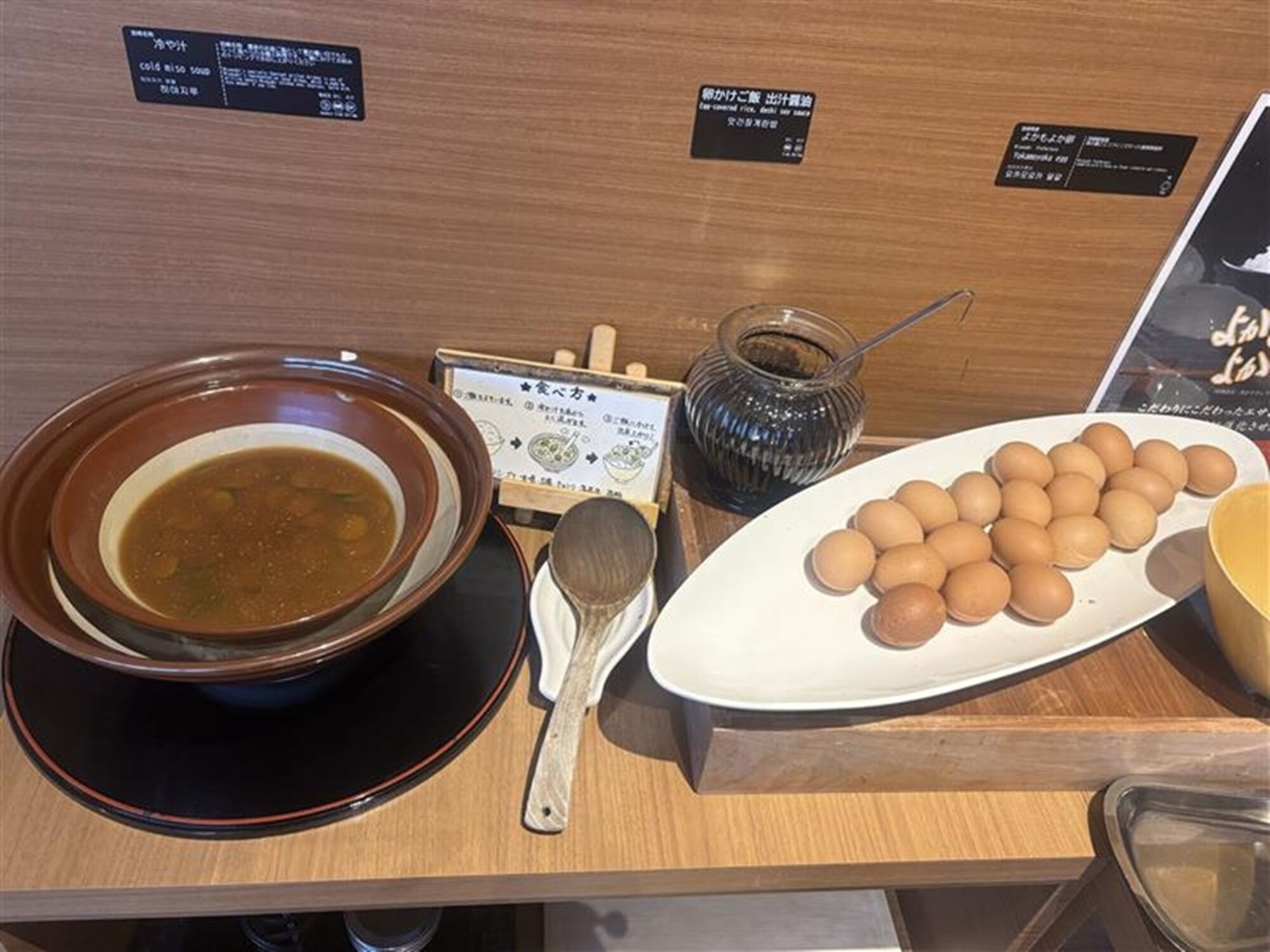 A table with a bowl of soup, a plate of eggs, a jar of pickles, and a wooden spoon.