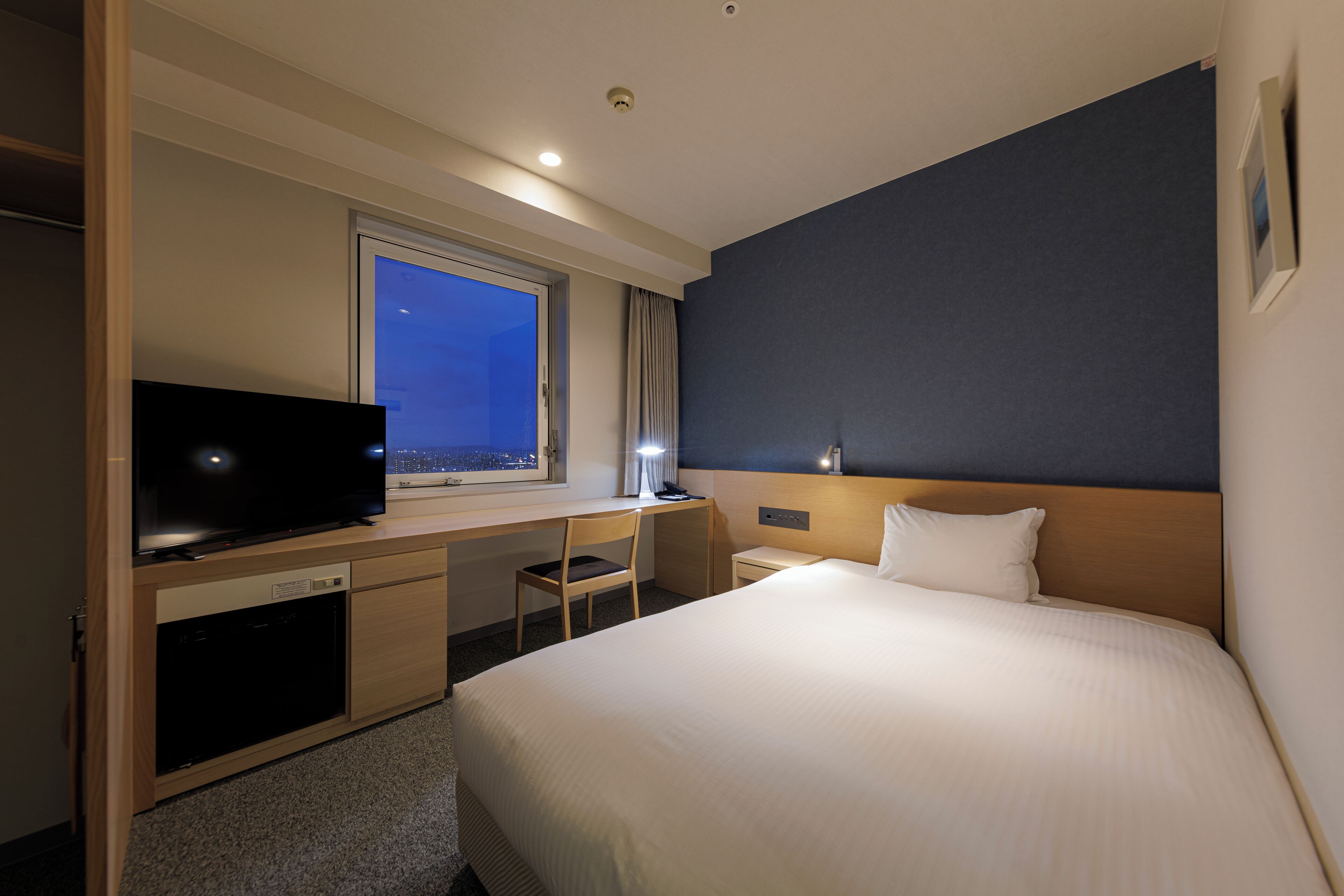 A hotel room with a bed, a desk with a television, a chair, a nightstand, and a window with a city view.
