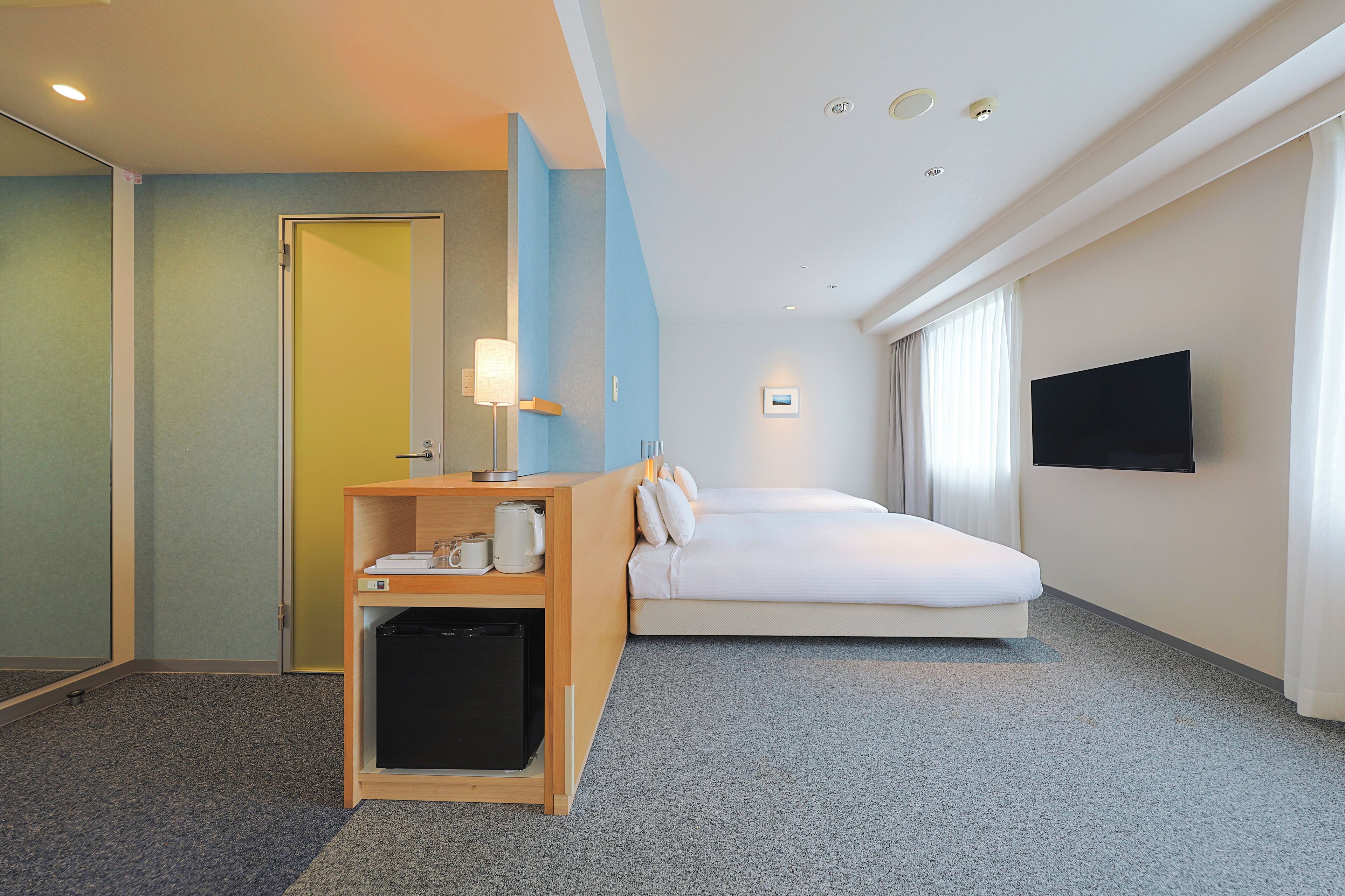 Una habitación de hotel con una cama, un escritorio con lámpara, un televisor en la pared y un pequeño armario.