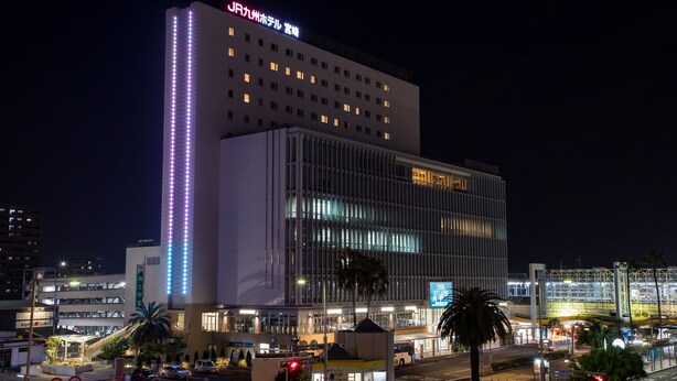 A modern hotel with a prominent vertical light strip on the facade, a large glass entrance, and a well-lit parking area. - JR KYUSHU HOTEL Miyazaki (Miyazaki)