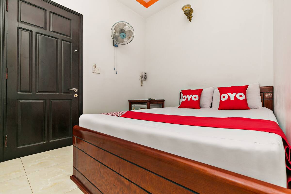 Une chambre avec un grand lit, une tête de lit en bois, une porte sombre, un ventilateur fixé au mur et une petite table de chevet.