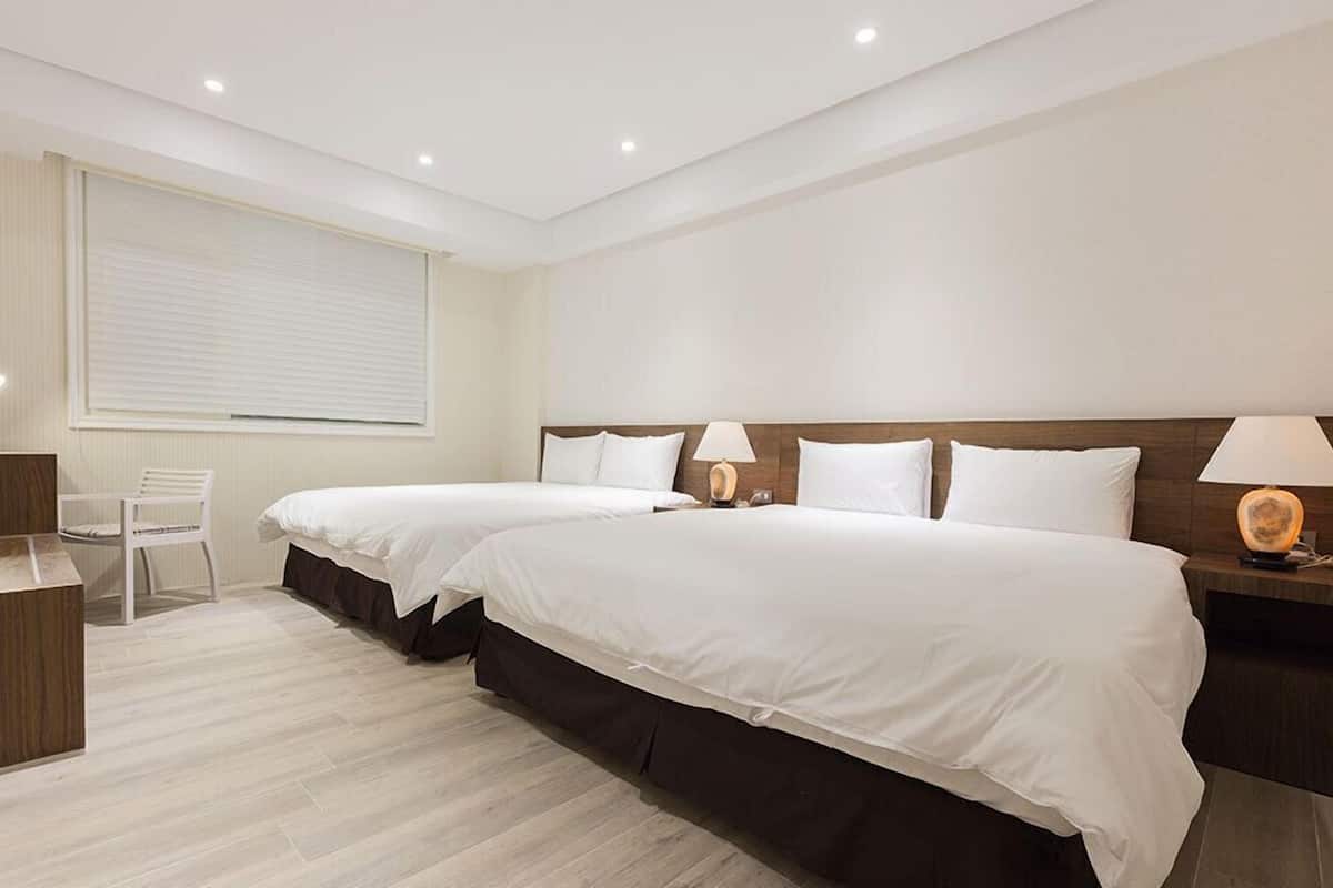 A hotel room with two beds, a desk, a chair, and a window with blinds.
