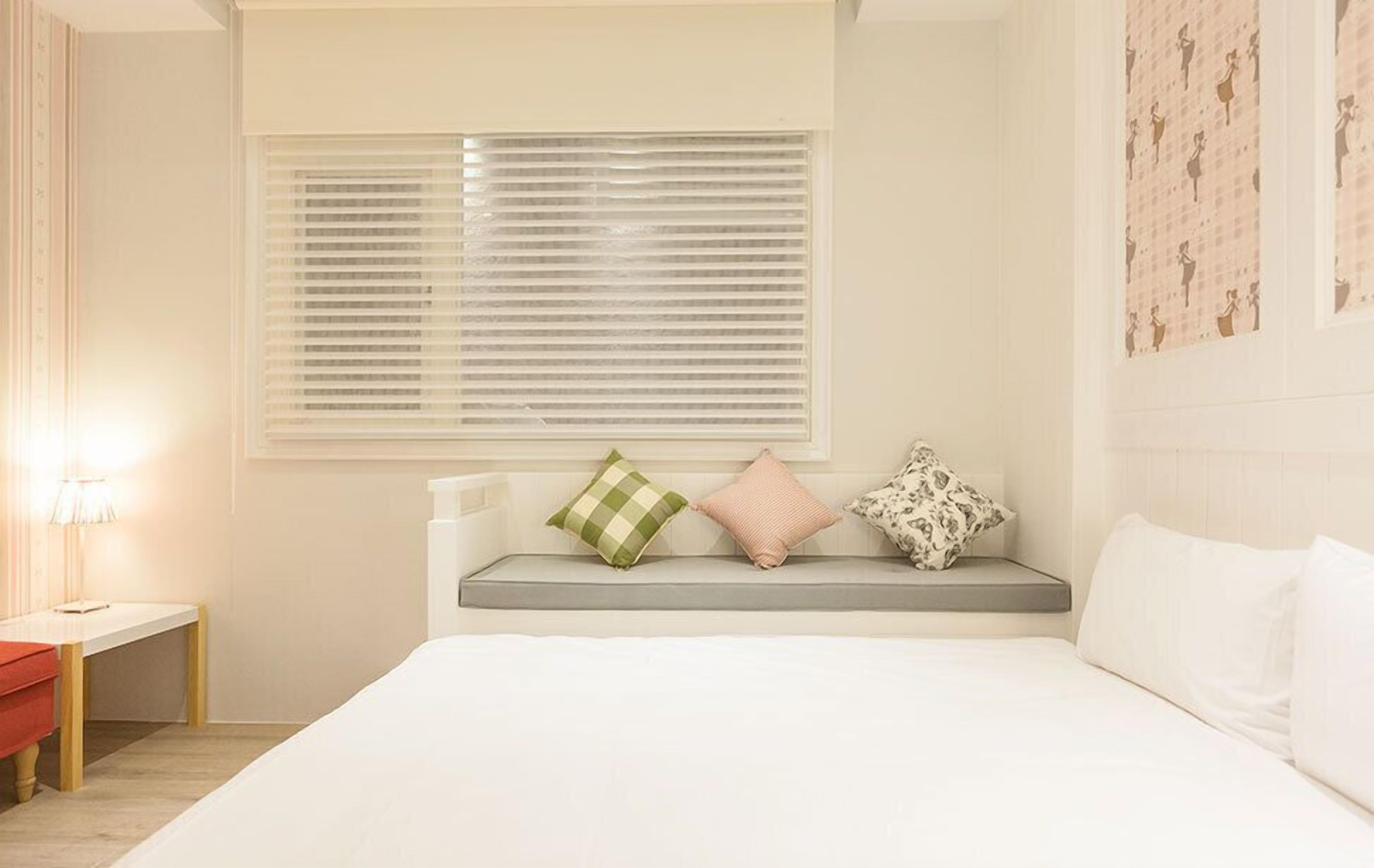 A bedroom with a bed, a bench with pillows, a window with blinds, and a wall with a pattern.