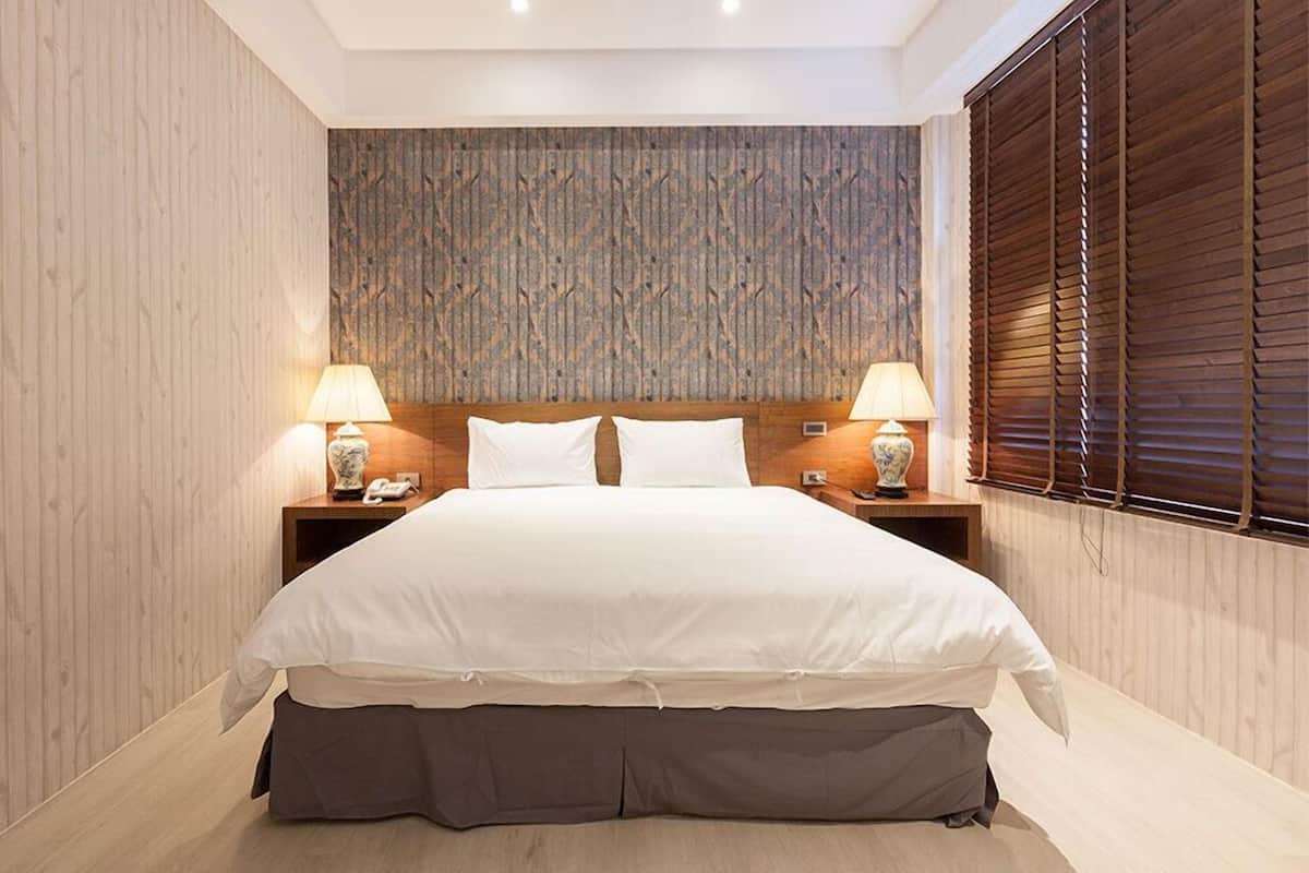 A modern bedroom with a large bed, wooden headboard, two bedside lamps, and a window with blinds.