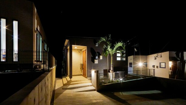 Un edificio moderno con una entrada bien iluminada, un pequeño jardín con una palmera y una escalera visible que conduce a la entrada.