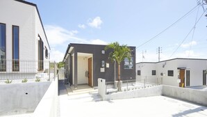 Un edificio residencial moderno con techo plano, un camino de hormigón y una palmera pequeña.
