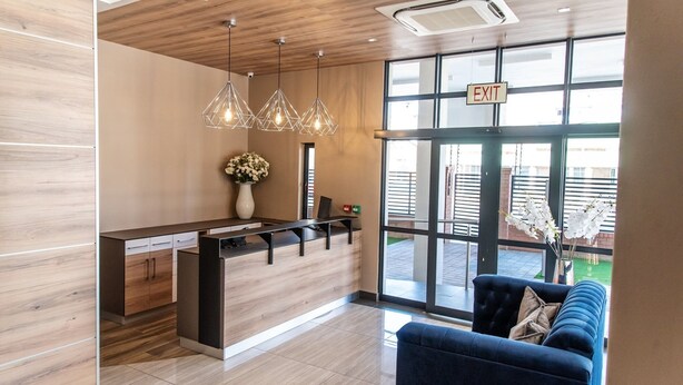 A modern reception area with a wooden reception desk, pendant lighting, a vase with flowers, and a blue sofa. - Loch Logan Hotel (Bloemfontein)