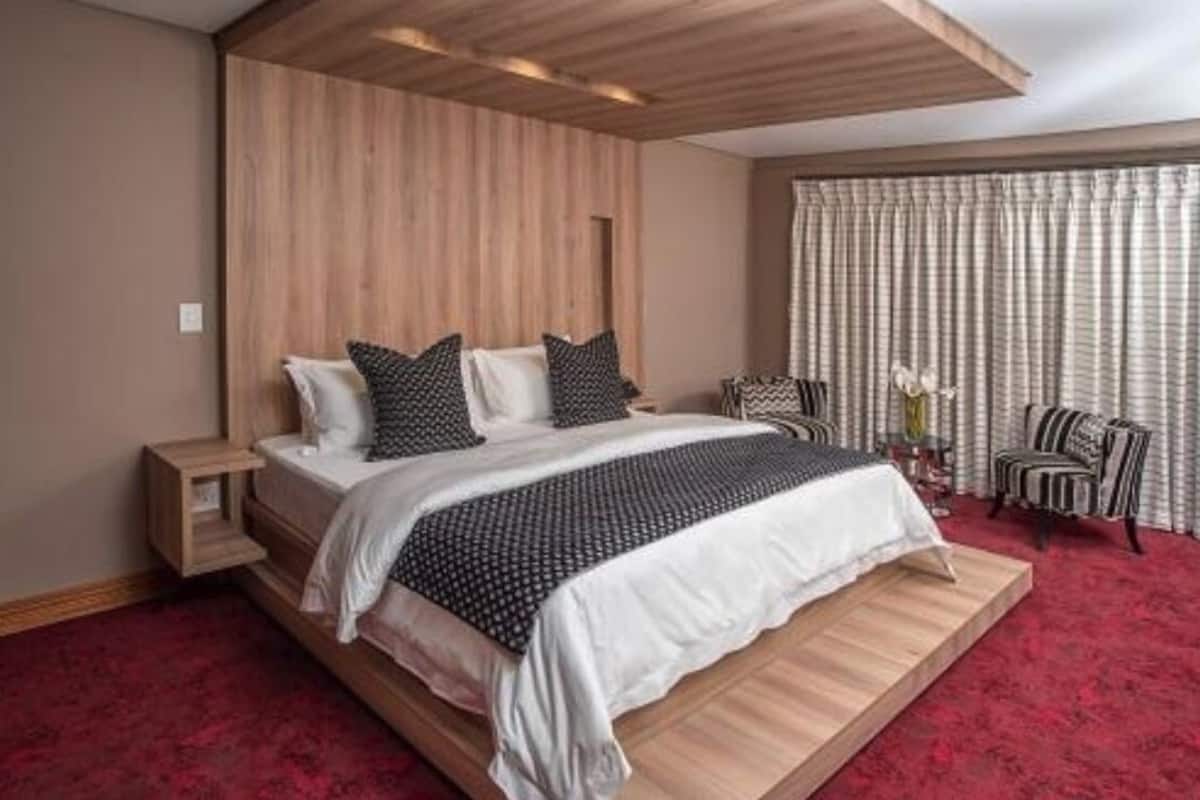 Ein modernes Schlafzimmer mit einem hölzernen Kopfteil, einem Bett mit weißer und gemusterter Bettwäsche, einem roten Teppich, einem Nachttisch und einem Stuhl mit gestreiften Kissen.