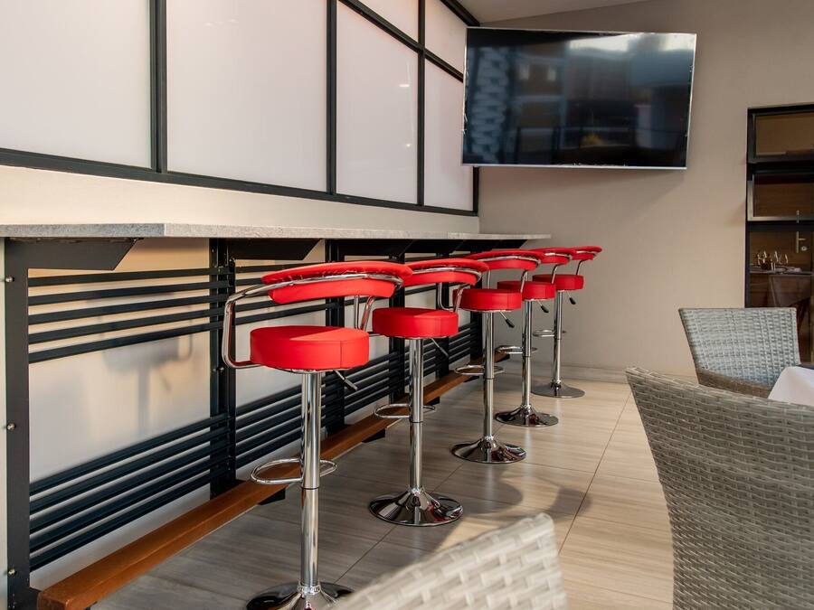 A row of red bar stools with chrome bases, a flat-screen TV mounted on the wall, and a wicker chair to the right.
