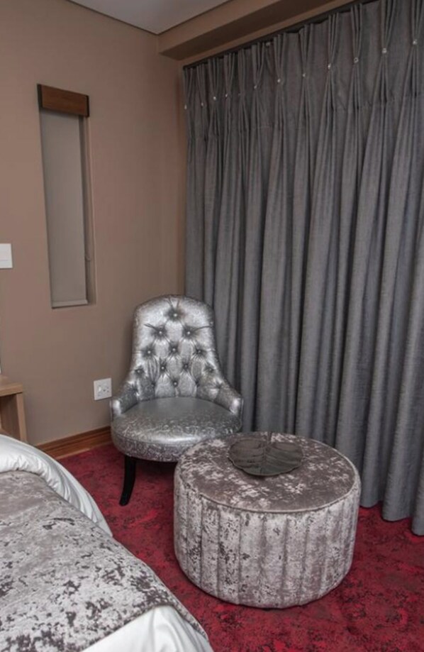 Une chambre d’hôtel avec un fauteuil à capitons, un pouf et un grand lit surmonté d’une moquette rouge.