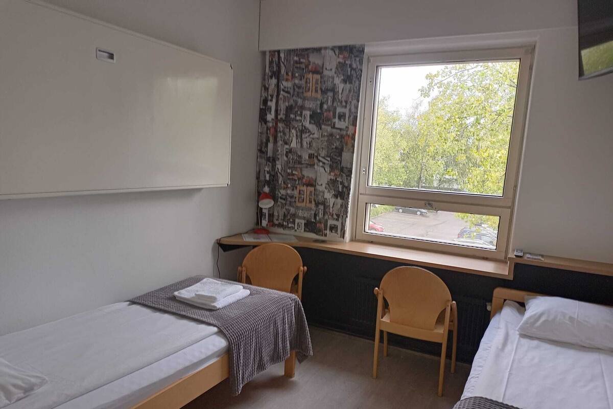 A dormitory room with two single beds, a desk with a chair, a window with a view of trees, and a wall with a collage of pictures.
