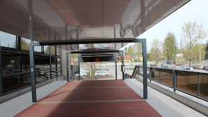 Un pasillo cubierto que conduce a un estacionamiento, con alfombra roja, barandillas metálicas y vistas a coches aparcados y árboles.