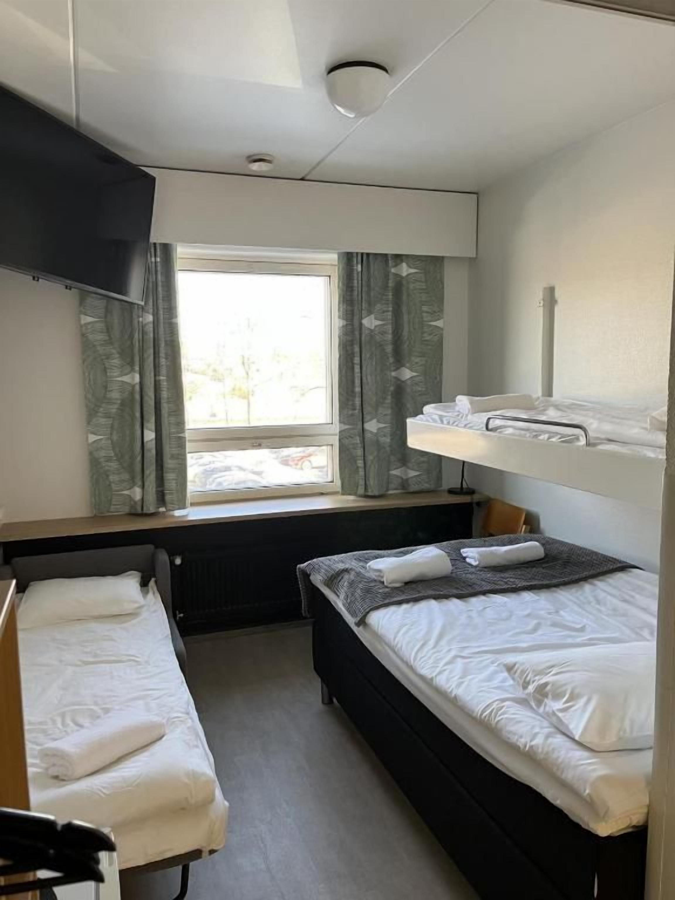 A compact hotel room with two beds, a window with curtains, a flat-screen TV, and a radiator.