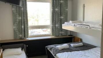 A compact hotel room with two beds, a window with curtains, a flat-screen TV, and a radiator.