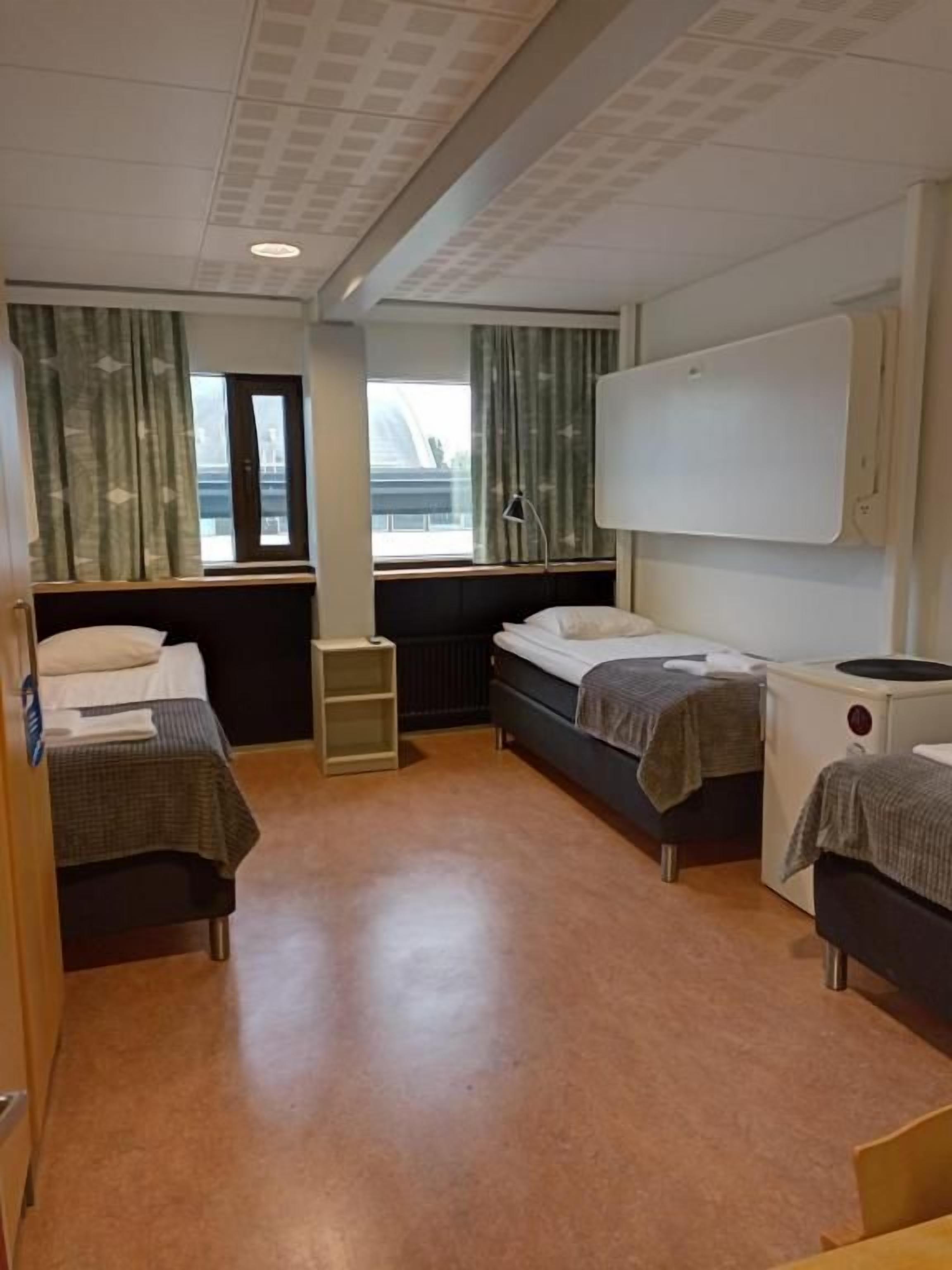 A compact hotel room with two beds, a small table, a chair, a window with curtains, and a ceiling with a grid pattern.