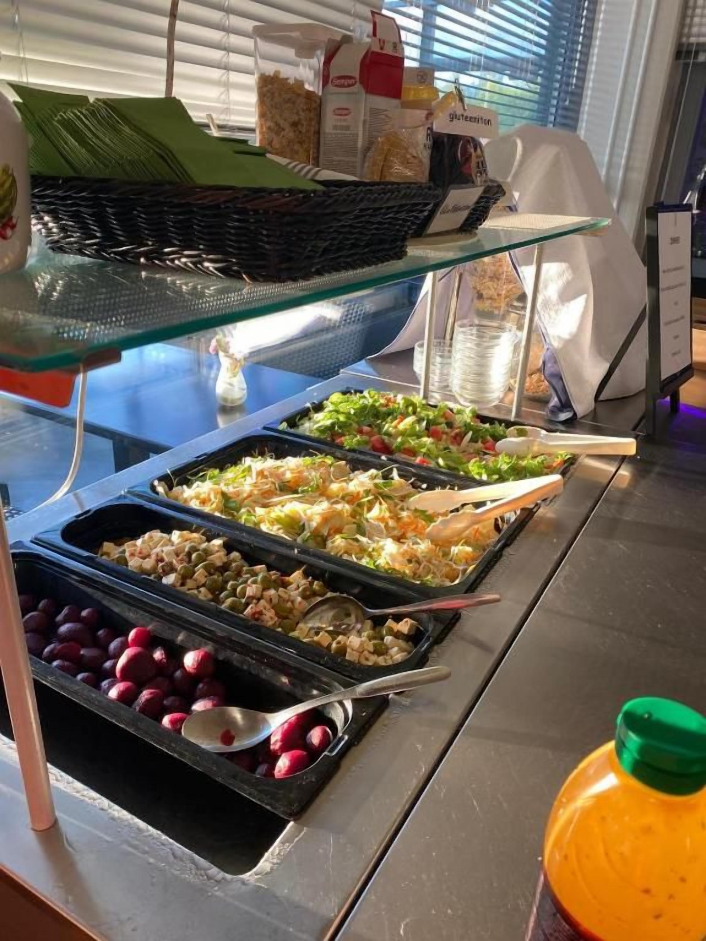 Monipuolinen buffet, jossa on tarjolla erilaisia ruokia, kuten salaatteja, hedelmiä ja juoma-automaatti.