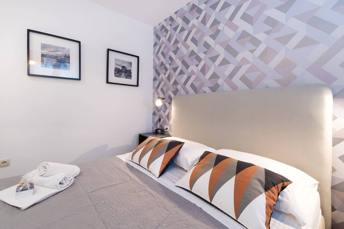 A hotel room with a bed, two framed pictures on the wall, a geometric-patterned wallpaper, and a pair of white towels on the floor.