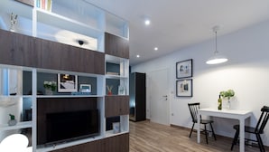 A modern living room with a flat-screen TV, wooden shelving, a dining area with a white table and black chairs, and a ceiling light. - Apartments Cingulice (Kastela)