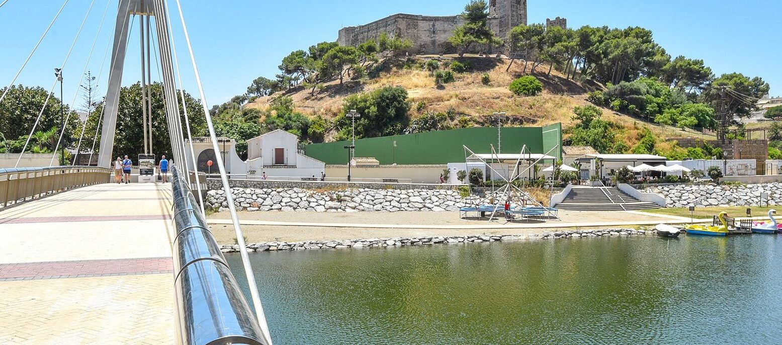 A bridge with a stone pathway leading to a body of water, a small building, and a hill with ruins in the background.