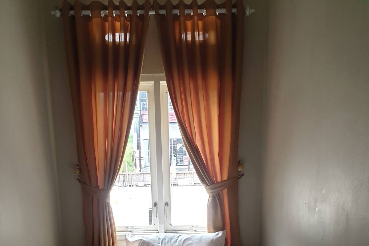 Ein kleines Zimmer mit einem Einzelbett, einem Fenster, Vorhängen und Außenblick.