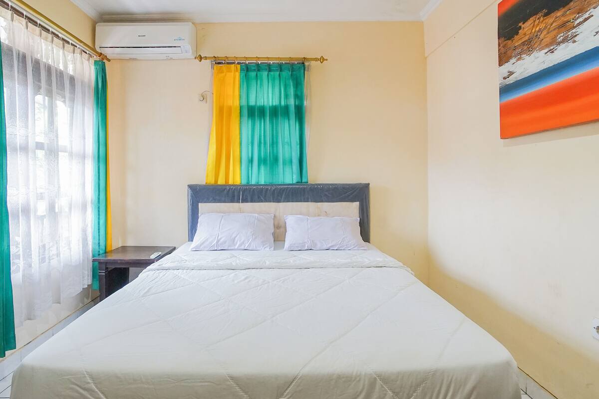 A hotel room with a bed, a nightstand, a window with curtains, an air conditioning unit, and a painting on the wall.