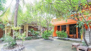 A courtyard with a paved floor, surrounded by various plants and trees, and a building with an orange facade. - Mahendra Beach Inn (Kuta)