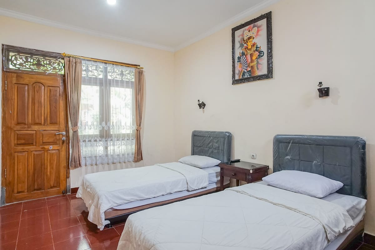 A room with two beds, a wooden door, a window with curtains, and a wall-mounted light fixture.