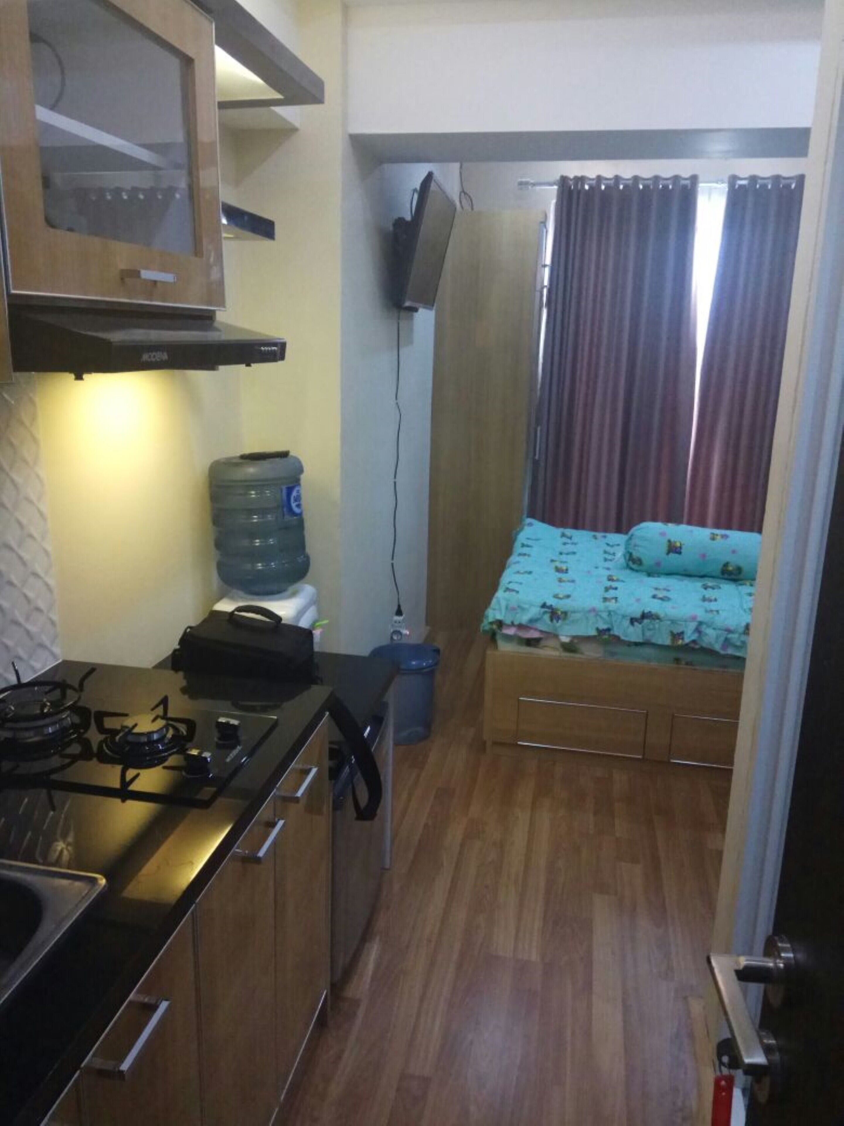 A compact kitchen with a stove, cabinets, and a water dispenser. A small bed with a blue patterned bedspread is visible in the background.